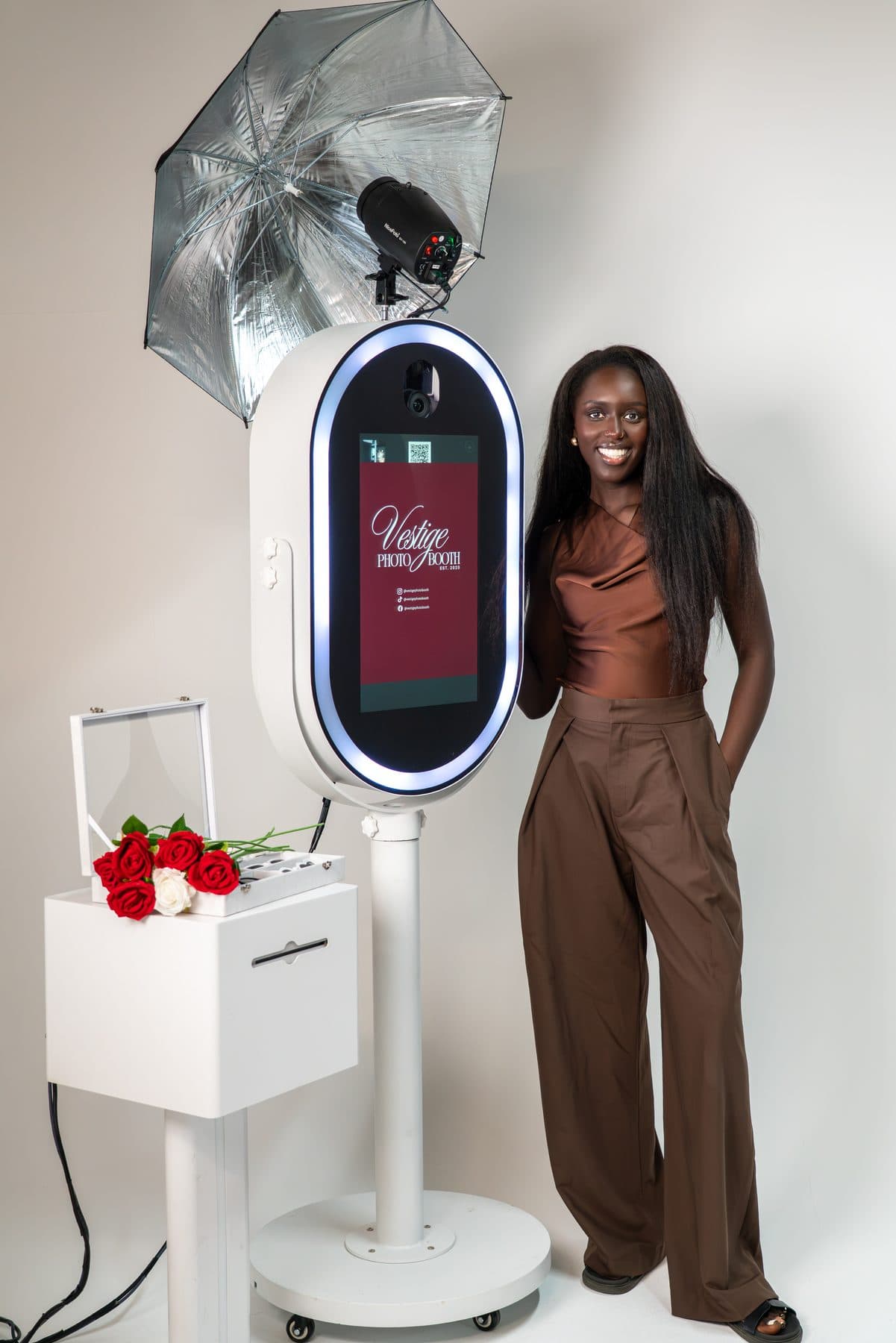Studio branding portrait of a woman in a brown satin top and wide trousers, smiling beside a Vestige Photo Booth with a box of red roses.